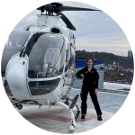 Caroline Levin in a flight suit smiling in front of a helicopter, standing confidently with hand on hip.