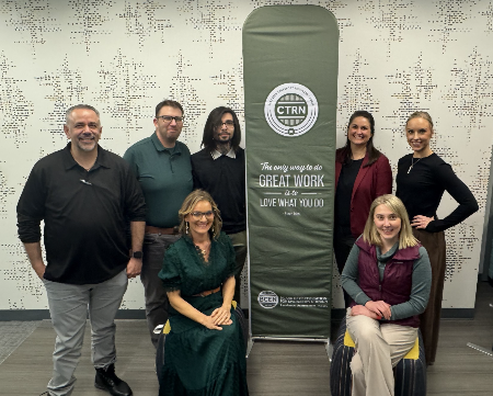 Seven people pose indoors around a green banner that reads "CTRN" and "The only way to do great work is to love what you do." Five are standing and two are seated in front.
