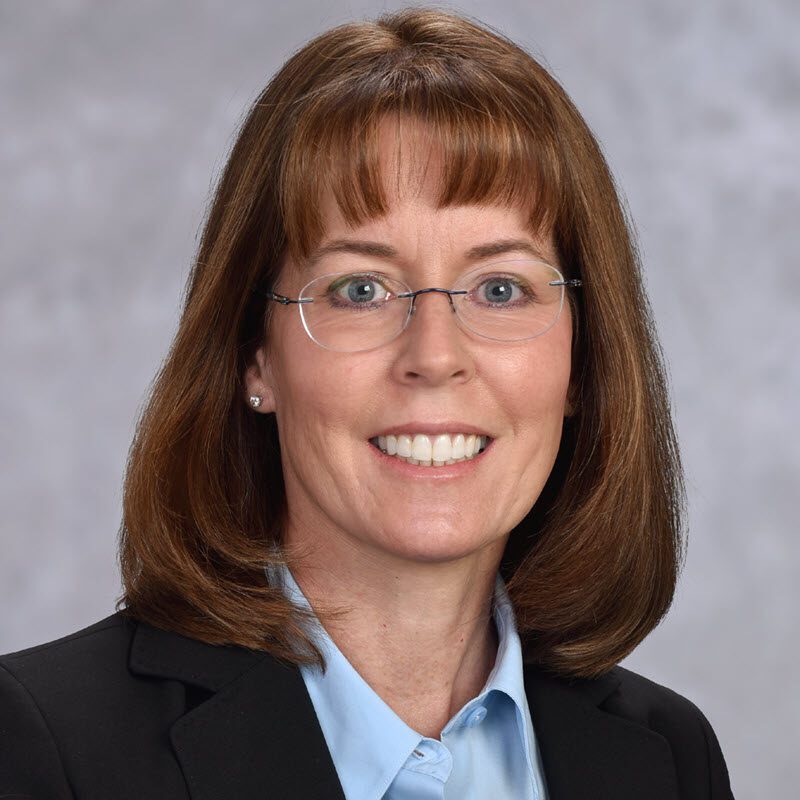 Woman with straight brown hair, wearing glasses, a black blazer, and a light blue collared shirt, smiling in front of a neutral gray background.
