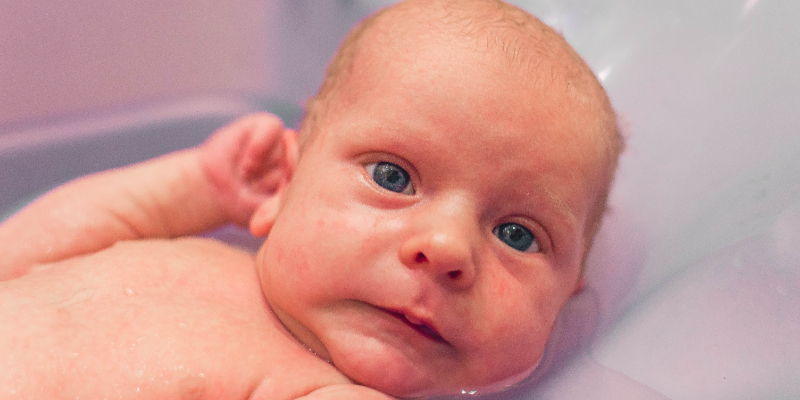 A newborn baby with blue eyes lies in a bath, supported by an adult hand, with a neutral facial expression.