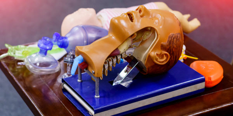 A medical airway training manikin head with an open mouth is displayed on a blue binder, surrounded by various airway management tools.