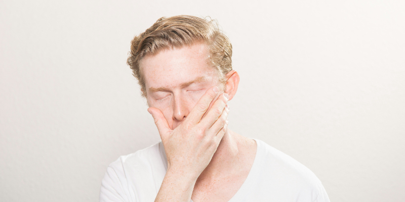 A man with closed eyes covers his mouth and lower face with one hand while standing against a plain, light background.