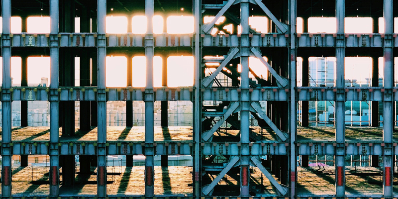 Steel framework of an unfinished building with sunlight streaming through the beams, casting long shadows on the ground below.