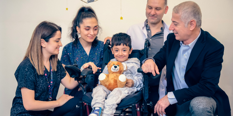 A young boy in a wheelchair holding a teddy bear is surrounded by three adults, all smiling and interacting with him indoors.