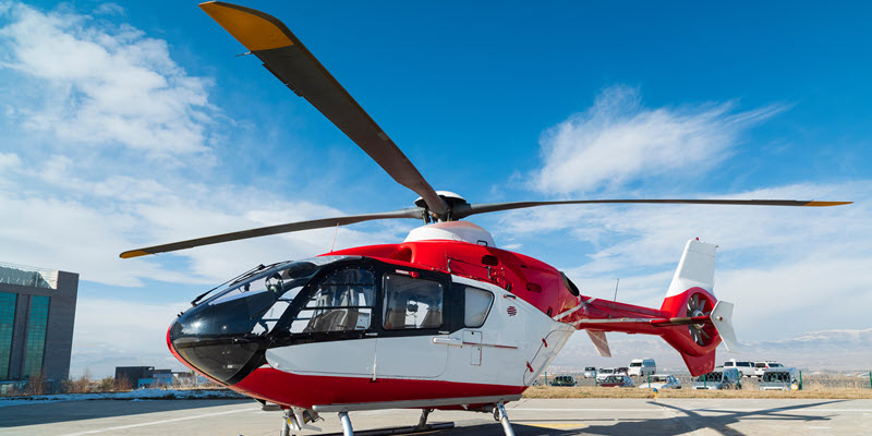 Red and white helicopter parked on a helipad under a clear blue sky, with a building and vehicles visible in the background.