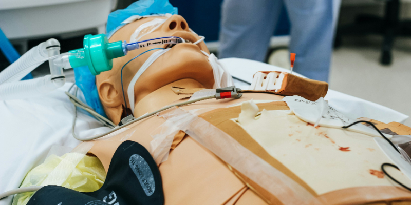 Medical training mannequin on a hospital bed with intubation tube, IV lines, and monitoring equipment attached, used for simulation practice.