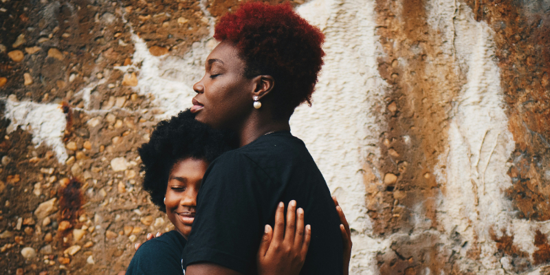Two people hug in front of a textured stone wall. One person has short red hair and closed eyes, while the other person smiles with their head against the other's chest.