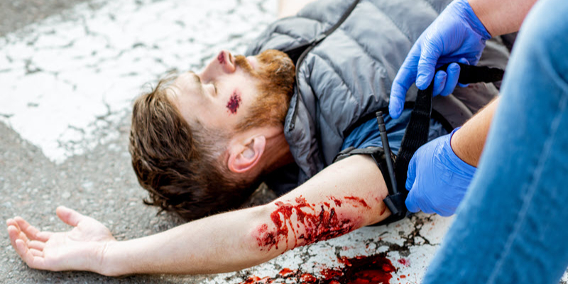 A person with visible injuries lies on the ground while someone wearing blue gloves applies a tourniquet to their bleeding arm.