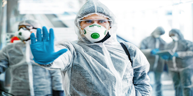 Person in full protective suit, mask, and gloves holds up hand to signal stop. Others in similar gear are in the background. Scene appears to be a biohazard or contamination area.