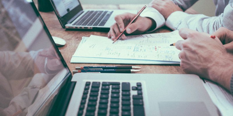 Two people working at a desk with laptops, reviewing documents and writing notes with pens.