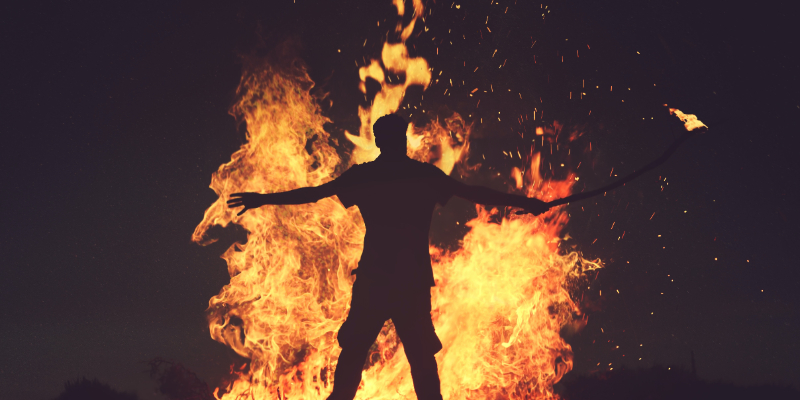 A person stands with arms outstretched in front of a large fire at night, holding a burning stick in one hand.