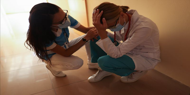 Two healthcare workers in scrubs and masks sit on the floor; one appears distressed with head in hand, while the other offers comfort.