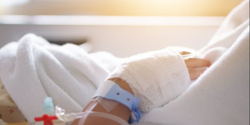 A close-up of a person's bandaged hand resting on a hospital bed, with an IV line and identification wristbands visible.
