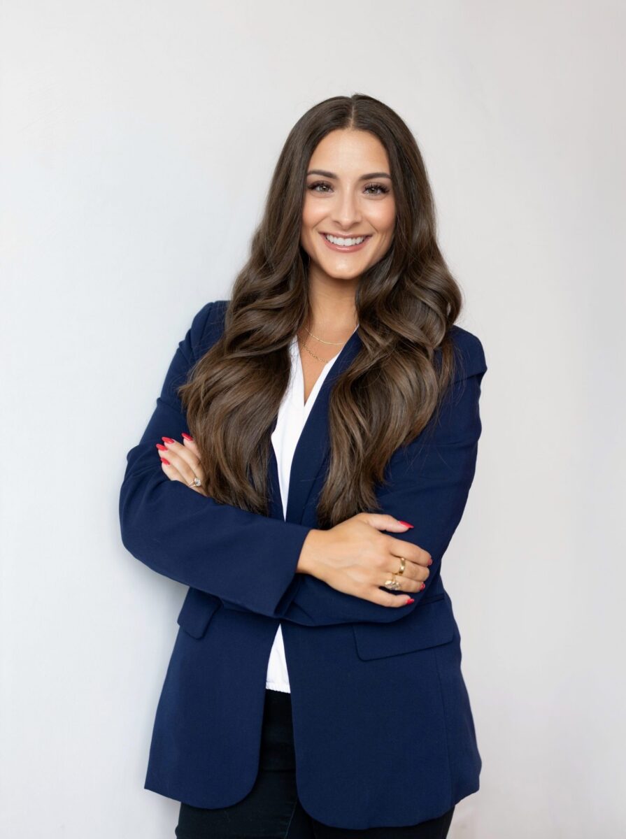 Woman with long brown hair wearing a navy blazer and white top, standing with arms crossed and smiling against a plain light background.