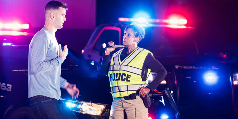 A police officer conducts a field sobriety test on a man at night with police cars and flashing lights in the background.