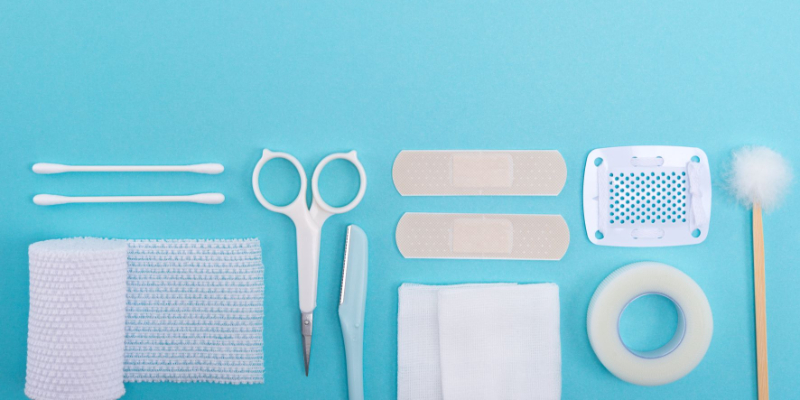 A set of medical supplies on a blue background, including gauze, cotton swabs, scissors, a scalpel, bandages, adhesive tape, and a cotton-tipped stick.