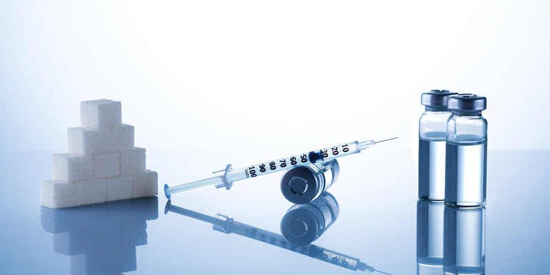 A syringe filled with liquid is placed between a stack of sugar cubes and two vials containing clear fluid, all on a reflective surface.