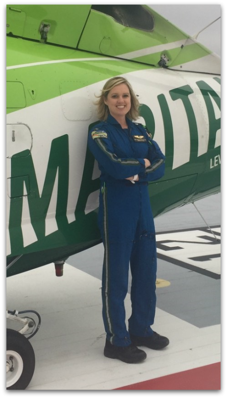 A woman in a blue flight suit stands smiling with arms crossed in front of a green and white helicopter.