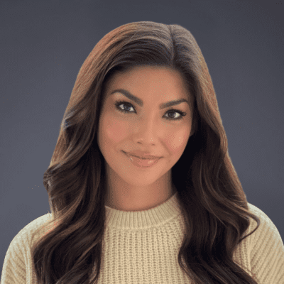Woman with long, wavy brown hair wearing a beige knit sweater, smiling softly, posed against a plain dark background.