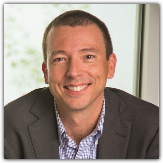 A man in a suit jacket and checkered shirt smiles at the camera while sitting indoors near a window.