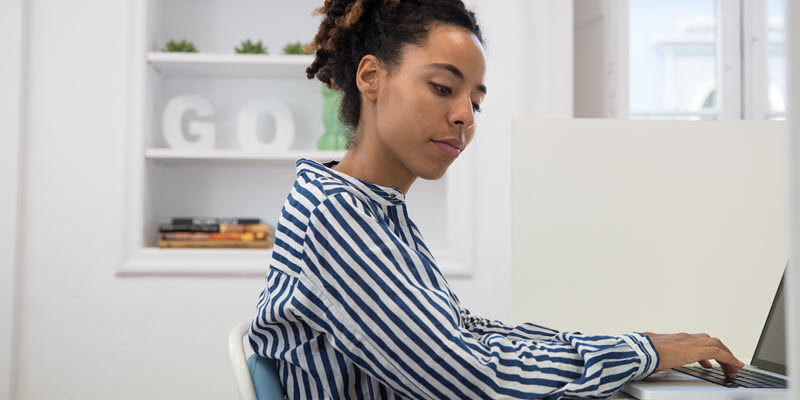 A person wearing a striped shirt sits at a desk and types on a laptop in a bright, modern room.
