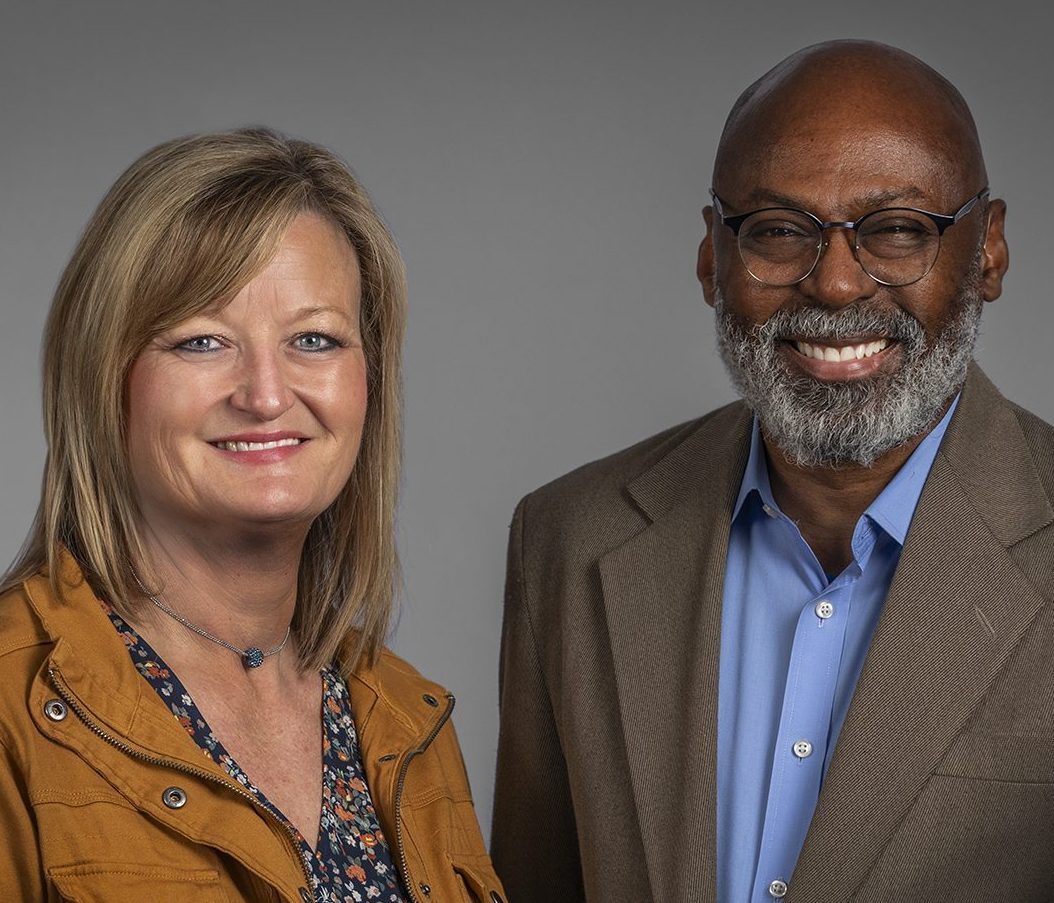 Two adults smile at the camera against a grey studio background. A woman in a tan jacket stands beside a bespectacled man in a brown blazer and blue shirt.