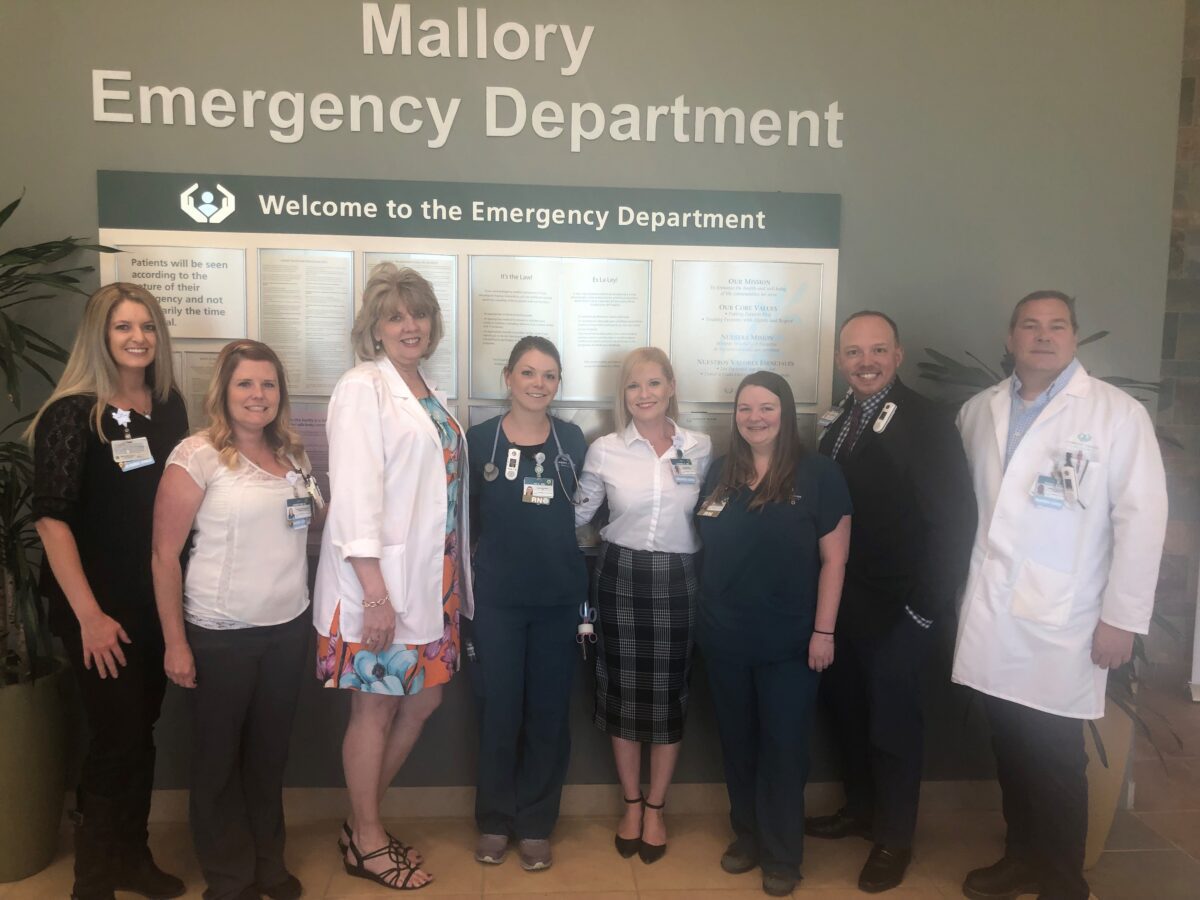 Eight hospital staff members stand in front of a sign reading "Mallory Emergency Department" and a welcome board inside a hospital.