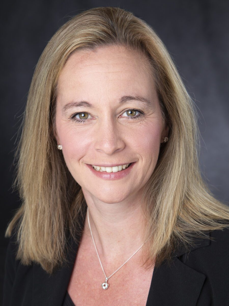 A woman with straight blonde hair and light skin wearing a black blazer and a necklace, smiling at the camera against a dark background.