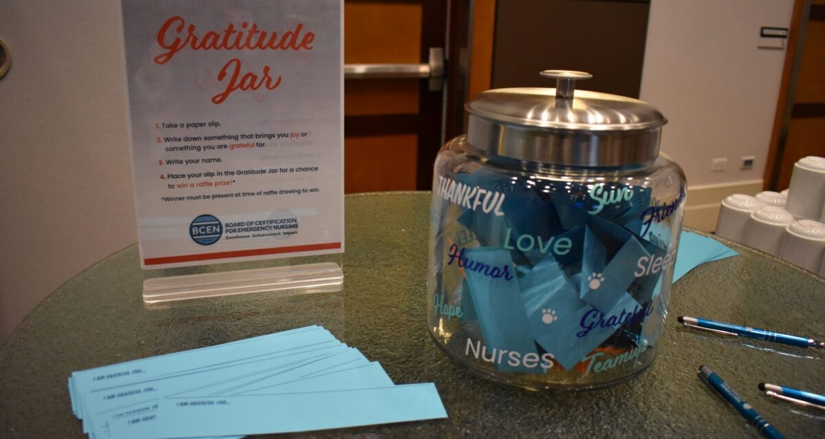 A glass jar labeled with words like "Thankful" and "Love" sits beside a sign titled "Gratitude Jar," blue paper slips, and pens on a table.