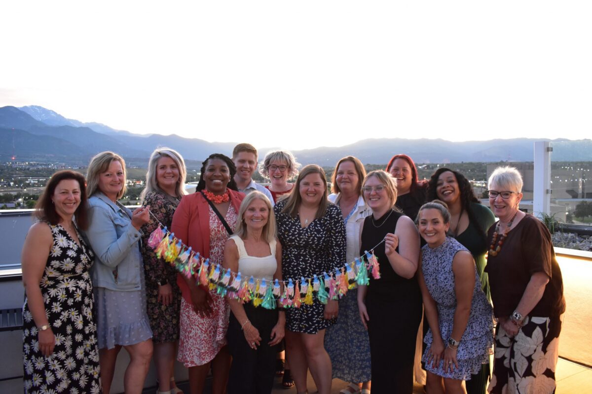 A group of people stand together outdoors at sunset, smiling and holding a colorful decorative banner, with mountains and a cityscape in the background.