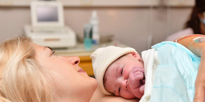 A newborn baby wearing a hat lies on the chest of a woman in a hospital setting shortly after birth.