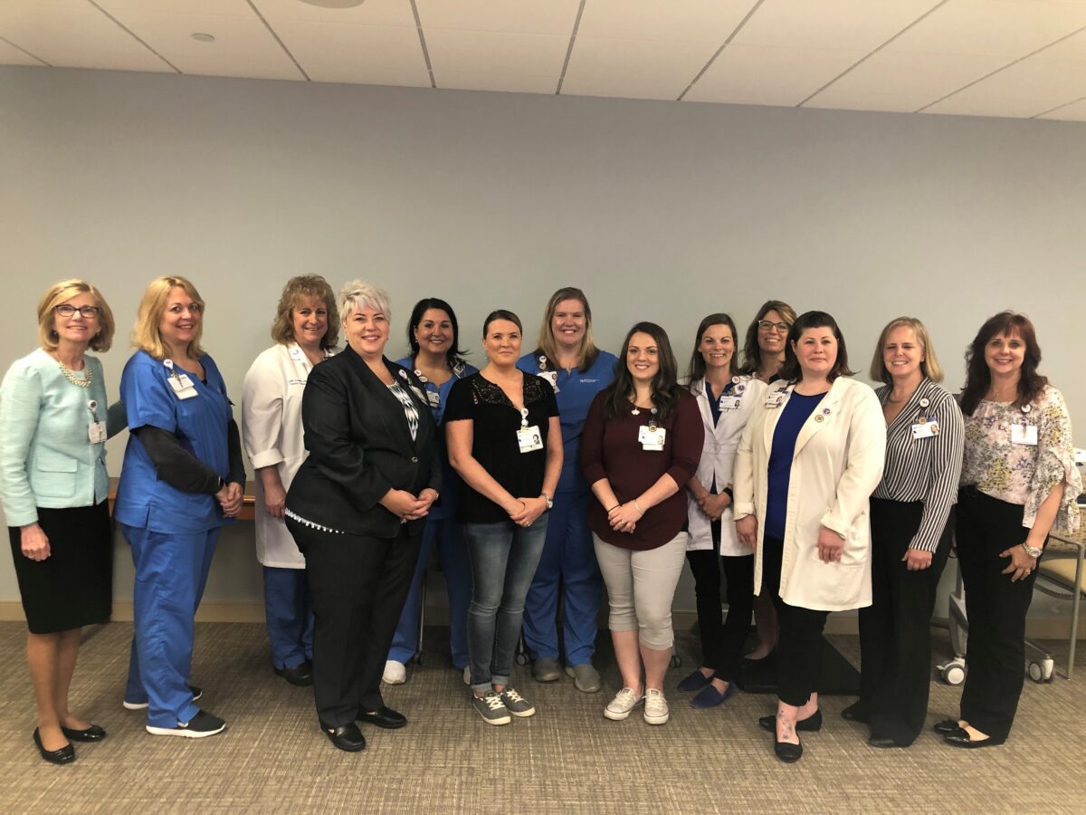 A group of thirteen women, including healthcare professionals in scrubs and lab coats, pose together indoors against a plain wall.