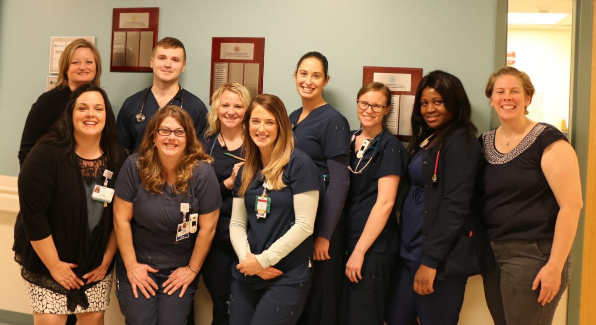 A group of ten healthcare professionals, some in scrubs and others in business attire, pose together and smile in a hallway with plaques on the wall behind them.