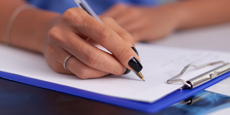 A person with black nail polish and a ring writes on a sheet of paper attached to a blue clipboard.