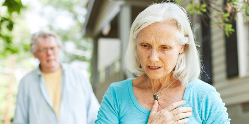An older woman in a blue shirt holds her chest with a concerned expression, while an older man stands in the background outside near a house.