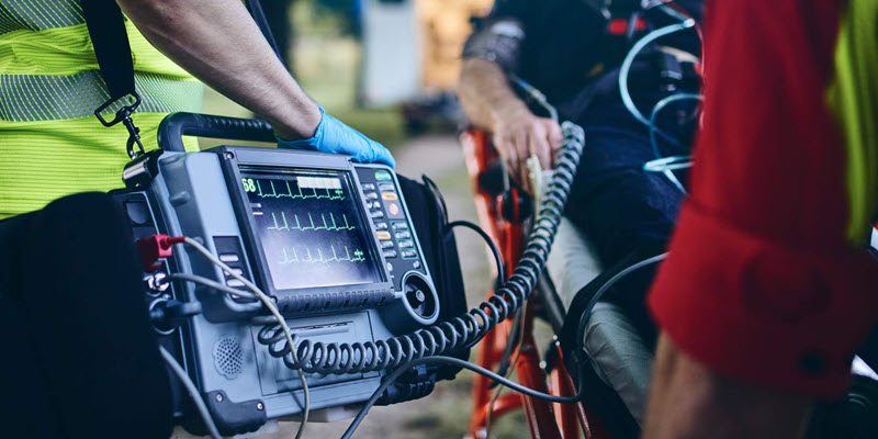 A medical professional operates a portable defibrillator, monitoring a patient’s vital signs on a stretcher during an emergency situation.