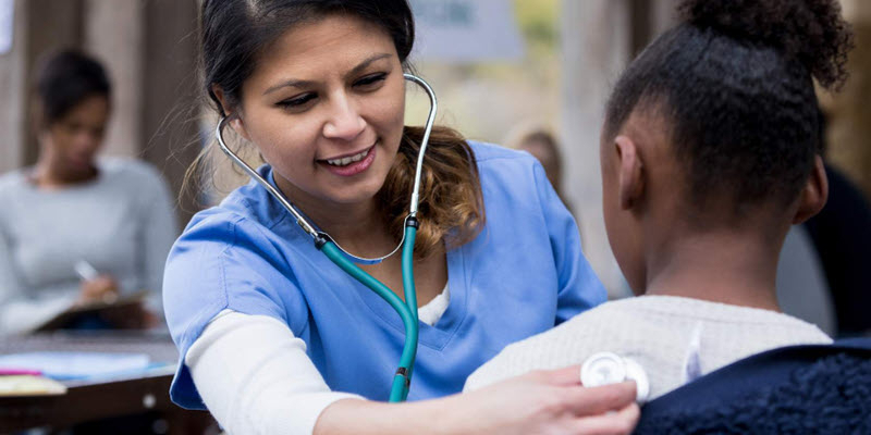 A healthcare worker in blue scrubs uses a stethoscope to listen to a young patient's back during an outdoor checkup.