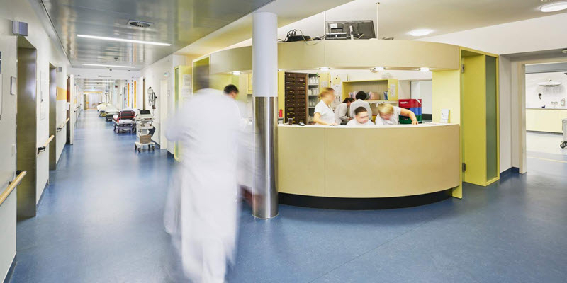 Hospital hallway with medical staff working behind a circular nurses' station and a staff member walking past in a white coat.