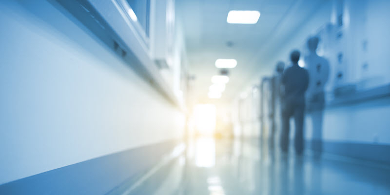 Blurred hospital corridor with two people standing near the wall and bright light shining at the end of the hallway.