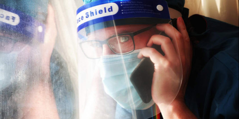 Person wearing glasses, a face mask, and a face shield talks on a phone while looking out a window.