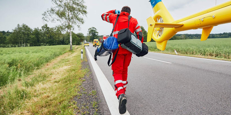 Paramedic in red uniform carries medical equipment toward an emergency scene on a rural road, with a yellow rescue helicopter and ambulance in the background.