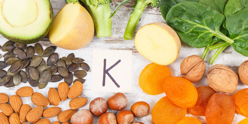 Assorted foods high in potassium, including potatoes, spinach, broccoli, nuts, seeds, and dried apricots, arranged around a card with the letter "K".