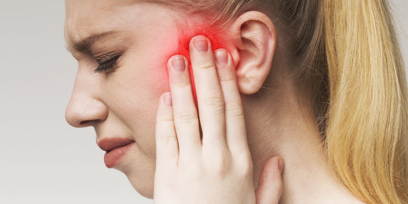 A woman touches her ear with a pained expression; the area around her ear is highlighted red, indicating ear pain or infection.