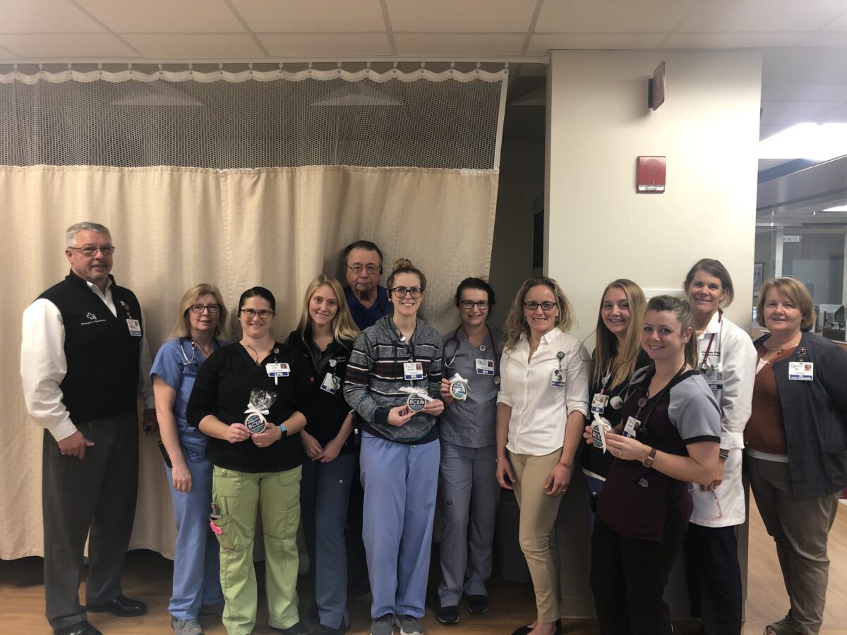 A group of healthcare workers, both men and women, stand indoors in a hospital setting, posing together and smiling for a group photo.