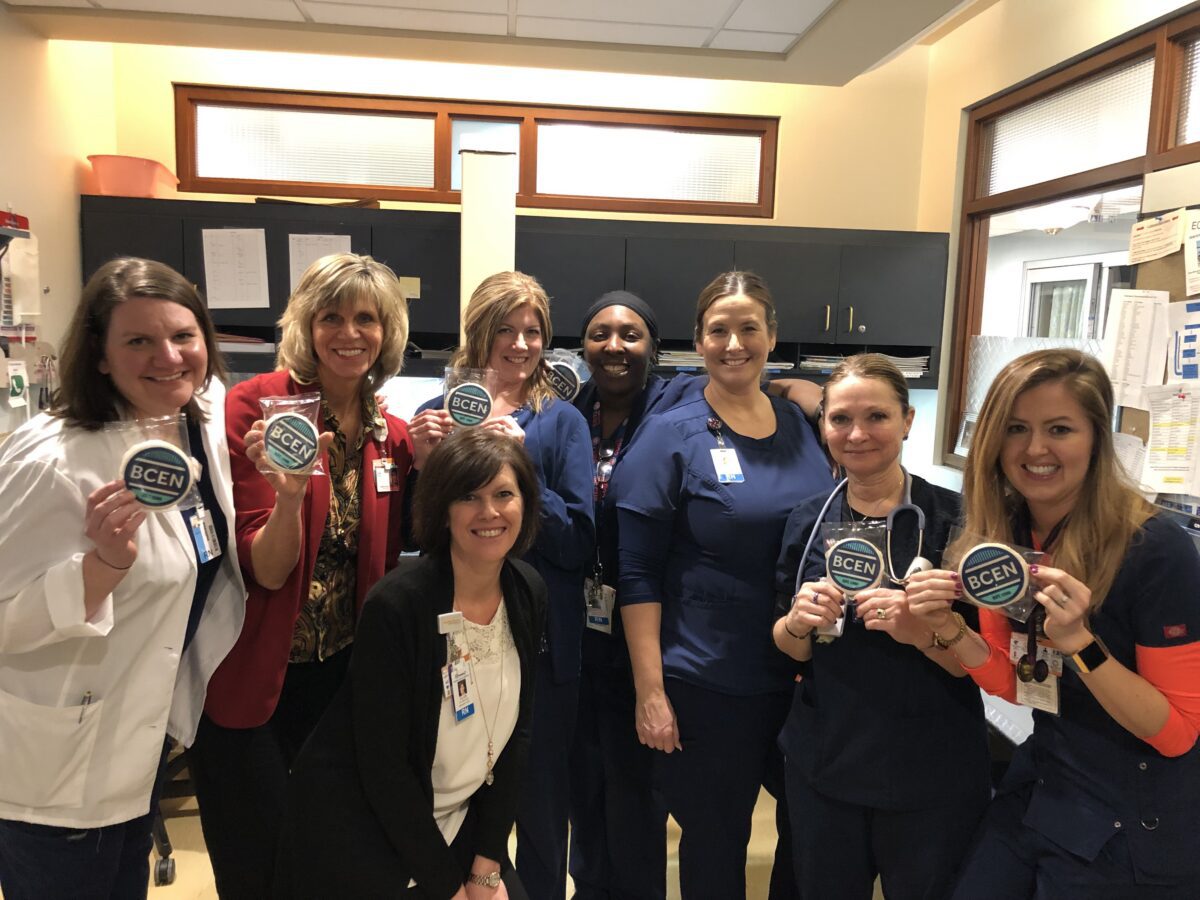 Eight women in medical attire stand together in a clinic, some holding circular "BCEN" badges and smiling at the camera.