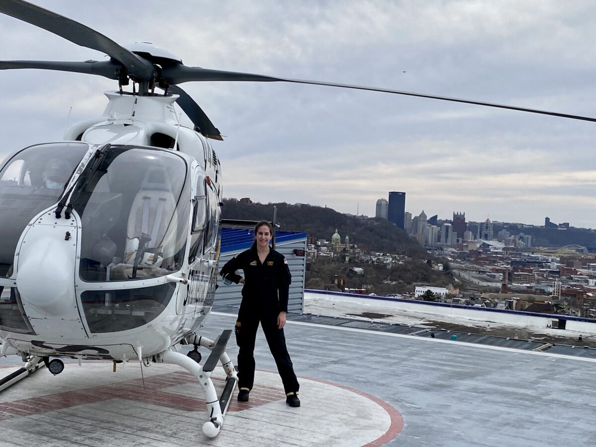 A person in a black flight suit stands next to a white helicopter on a rooftop helipad, with a city skyline and overcast sky in the background.