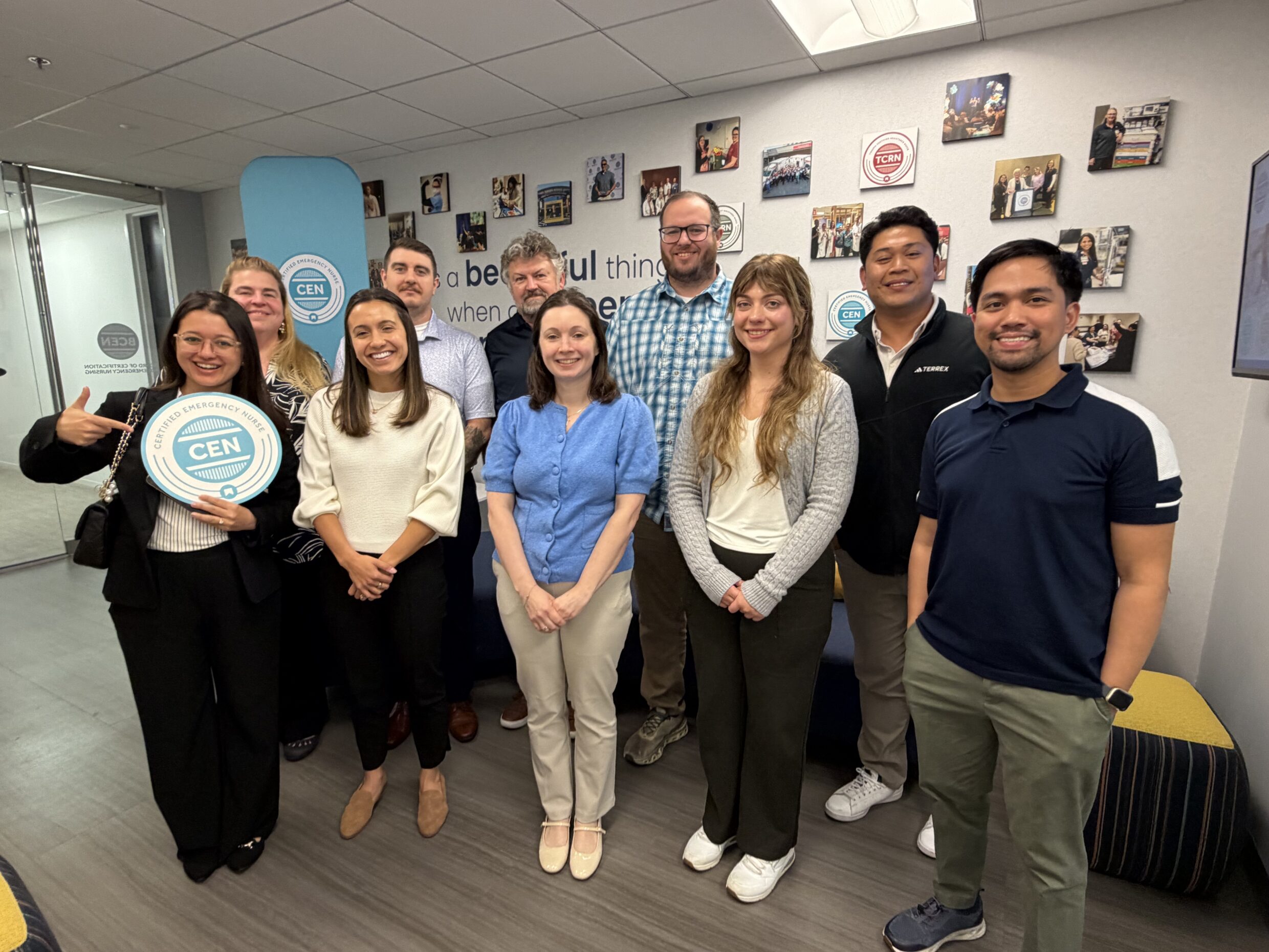 A group of ten people, the 2026 Certified Emergency Nurse (CEN) Passing Point Study Advisory Committee volunteer RNs, stand and smile in BCEN's office space, with one person holding a round "CEN" sign; photos and a quote decorate the wall behind them.