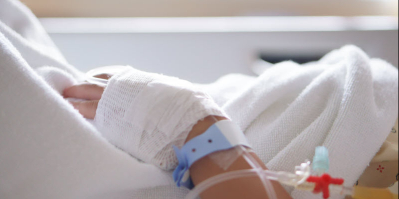 A close-up of a person's bandaged hand with a hospital wristband and an IV line, resting on a white blanket.