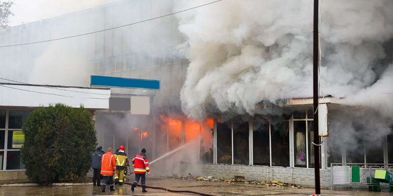 Firefighters spray water on a burning building with heavy smoke billowing out, while wearing safety gear and standing on wet ground.