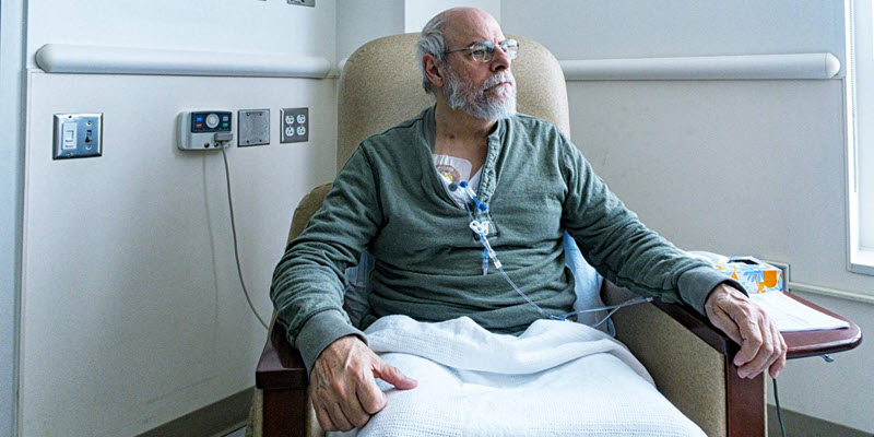 An older man sits in a hospital chair receiving intravenous treatment, with medical tubing attached to his chest and sunlight coming through the window.
